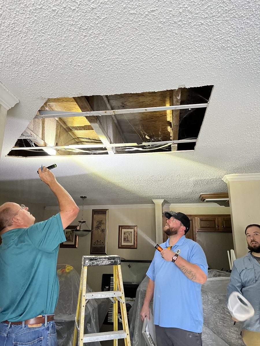 A hand shines a flashlight into an opening cut in a unit ceiling, exposing the floor framing above